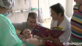 A l'hôpital de jour, une auxiliaire de puericulture distrait avec un livre une petite fille accompagnée de son papa, pendant un lavement de cathéter central.